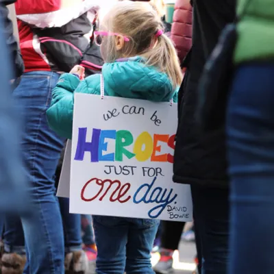 child supporting LGBTQ rights and gender transition at a rally with a sign that says "we can be heroes just for one day"